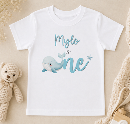 A white t-shirt with a blue whale graphic and the text "Mylo is one" printed on it, along with a teddy bear and other baby items.