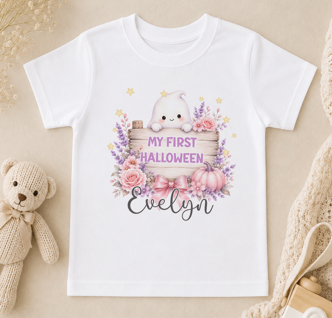 A white t-shirt with a Halloween-themed graphic design, including a ghost, flowers, pumpkins, and the text "My first Halloween Evelyn".