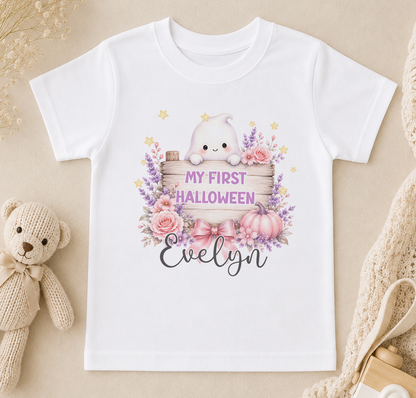 A white t-shirt with a Halloween-themed graphic design, including a ghost, flowers, pumpkins, and the text "My first Halloween Evelyn".