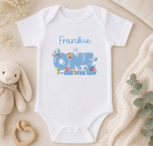 A white onesie with the text "Frankie is ONE - DER THE SEA" printed on it, along with various sea creatures such as fish and octopuses. The onesie is placed on a beige surface, surrounded by a stuffed animal, a wooden bead ring, and some greenery.