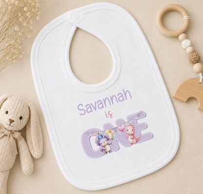 A white baby bib with the name "Savannah" and the number "1" printed on it, along with illustrations of a bunny and a dragon. The bib is placed on a surface with a stuffed animal and a wooden toy ring.