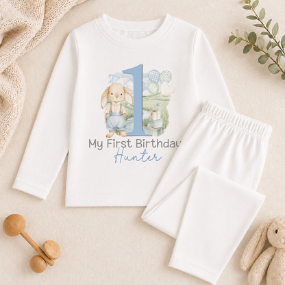 A white onesie with a blue number 1 and the text "My first birthday Hunter" printed on it, along with a pair of white pajama pants. The image also includes a stuffed animal and some other baby items.