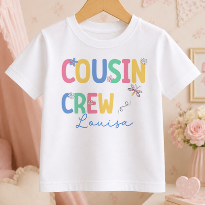 A white t-shirt with the text "COUSIN CREW Louisa" printed on it, along with colorful flowers and butterflies.