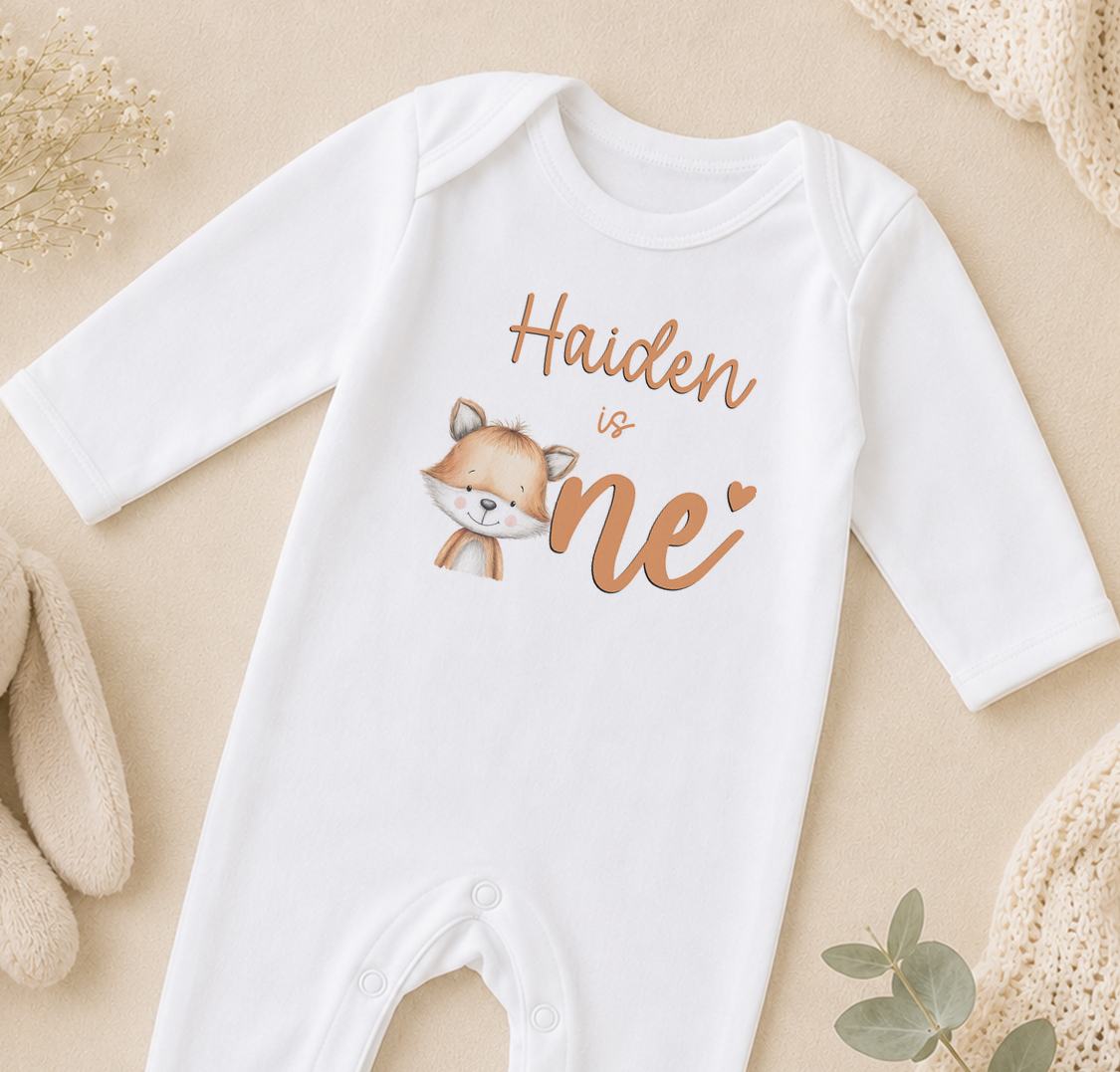 A white onesie with a cartoon fox and the text "Haiden is one" printed on it, along with a heart symbol.