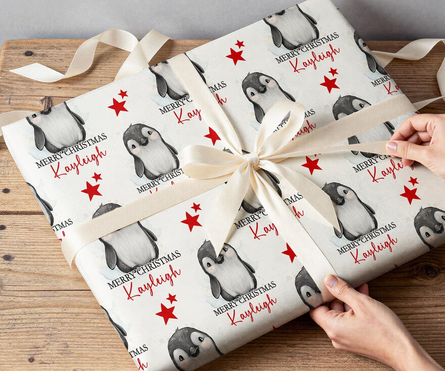 Hands wrapping a Christmas present in personalised penguin and red star wrapping paper with a white ribbon