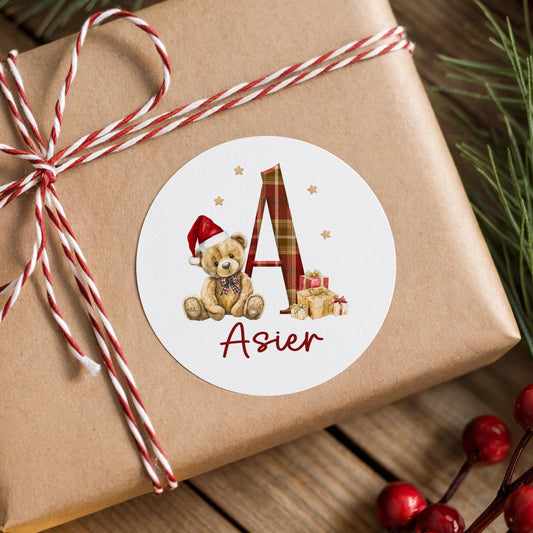 A round white label with a teddy bear wearing a Santa hat and the name "Asier" written on it, placed on a brown gift box wrapped with red and white striped twine.