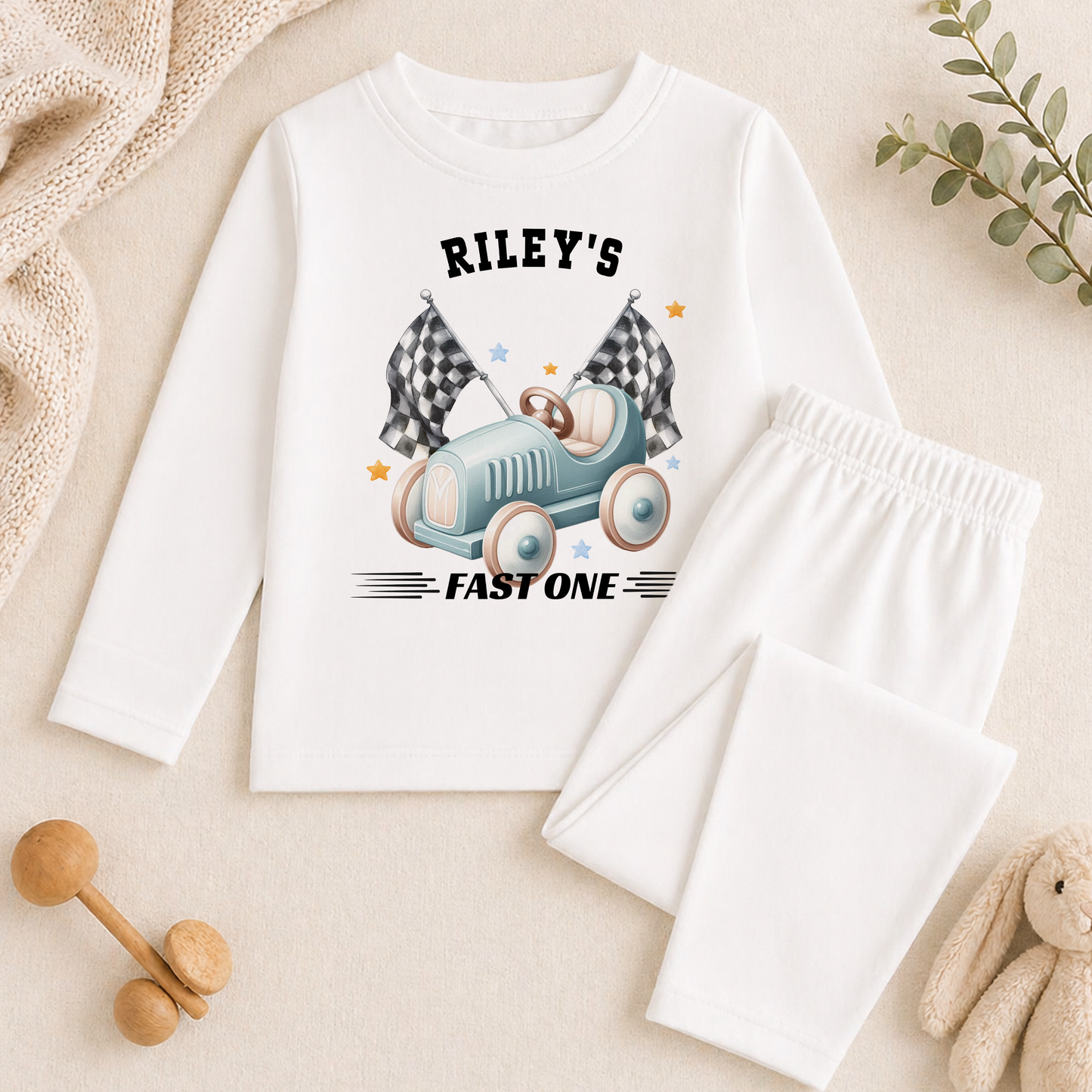 Long-sleeved top from the racing car birthday set, printed with a blue car, chequered flags and 'Riley's FAST ONE', styled with baby toys and a plant.