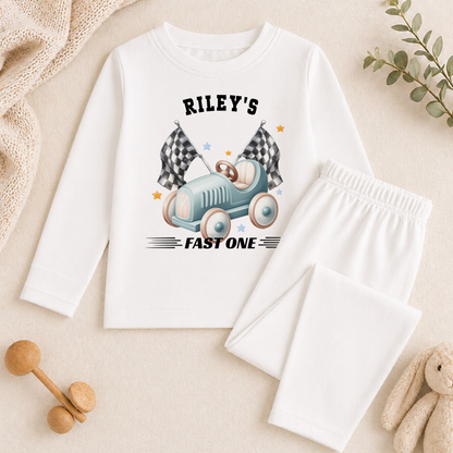 Long-sleeved top from the racing car birthday set, printed with a blue car, chequered flags and 'Riley's FAST ONE', styled with baby toys and a plant.