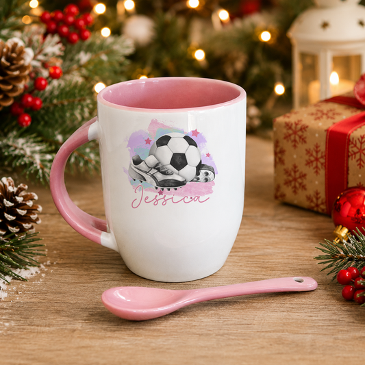 A white mug with a pink interior and a soccer ball design sits on a wooden surface, accompanied by a pink spoon. The mug is surrounded by Christmas decorations, including pine cones and ornaments.