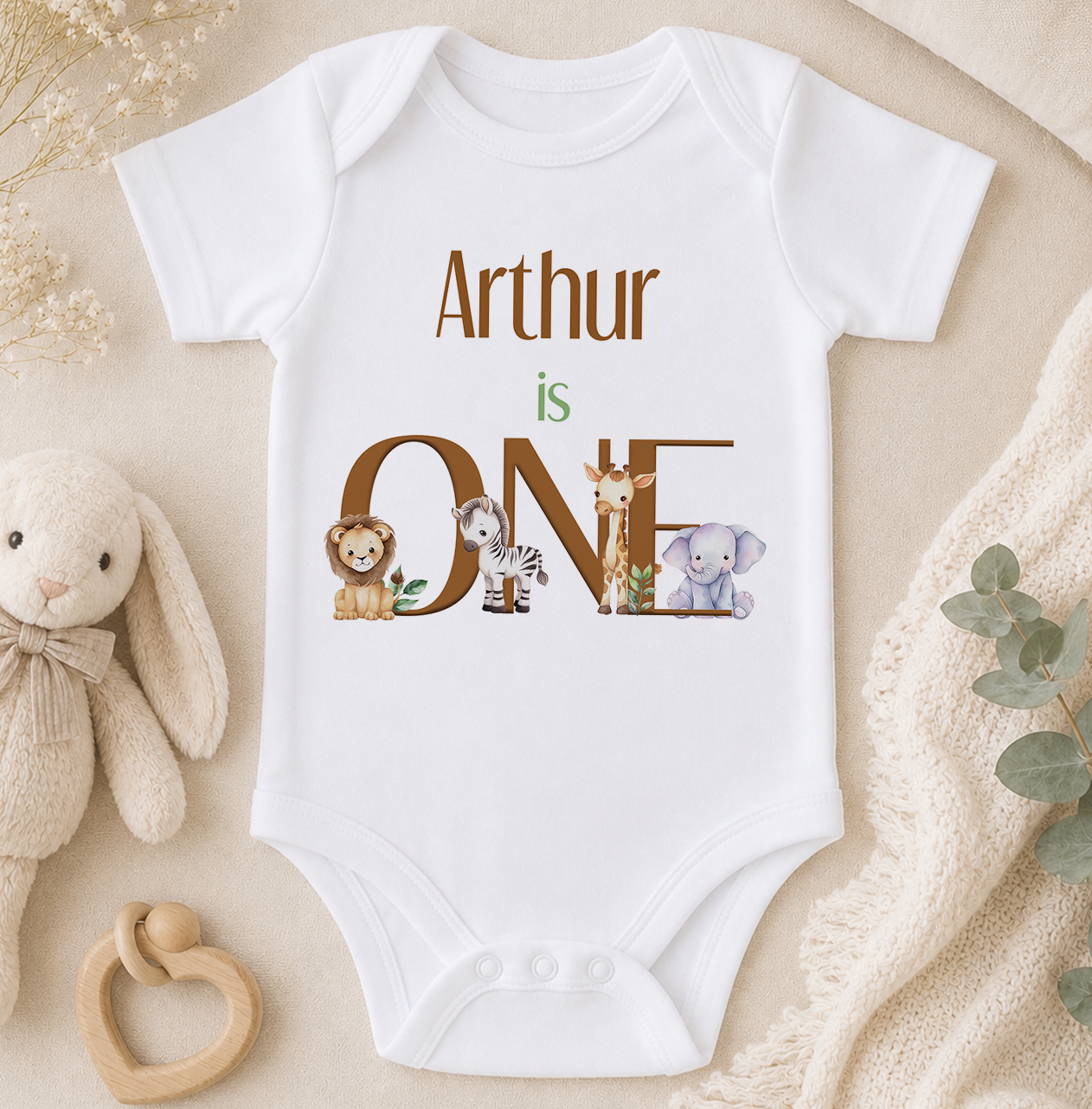 A white onesie with the text "Arthur is ONE" printed on it, surrounded by various animal illustrations, including a lion, giraffe, zebra, and elephant.