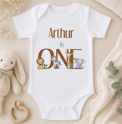 A white onesie with the text "Arthur is ONE" printed on it, surrounded by various animal illustrations, including a lion, giraffe, zebra, and elephant.