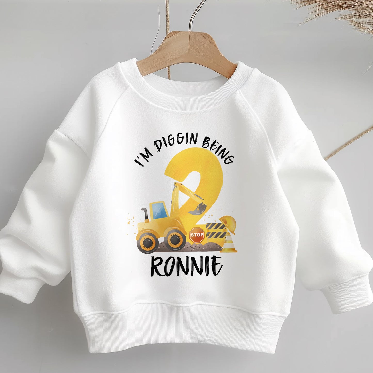 White children's sweatshirt on a hanger printed with a yellow digger construction birthday design reading I'M DIGGIN BEING