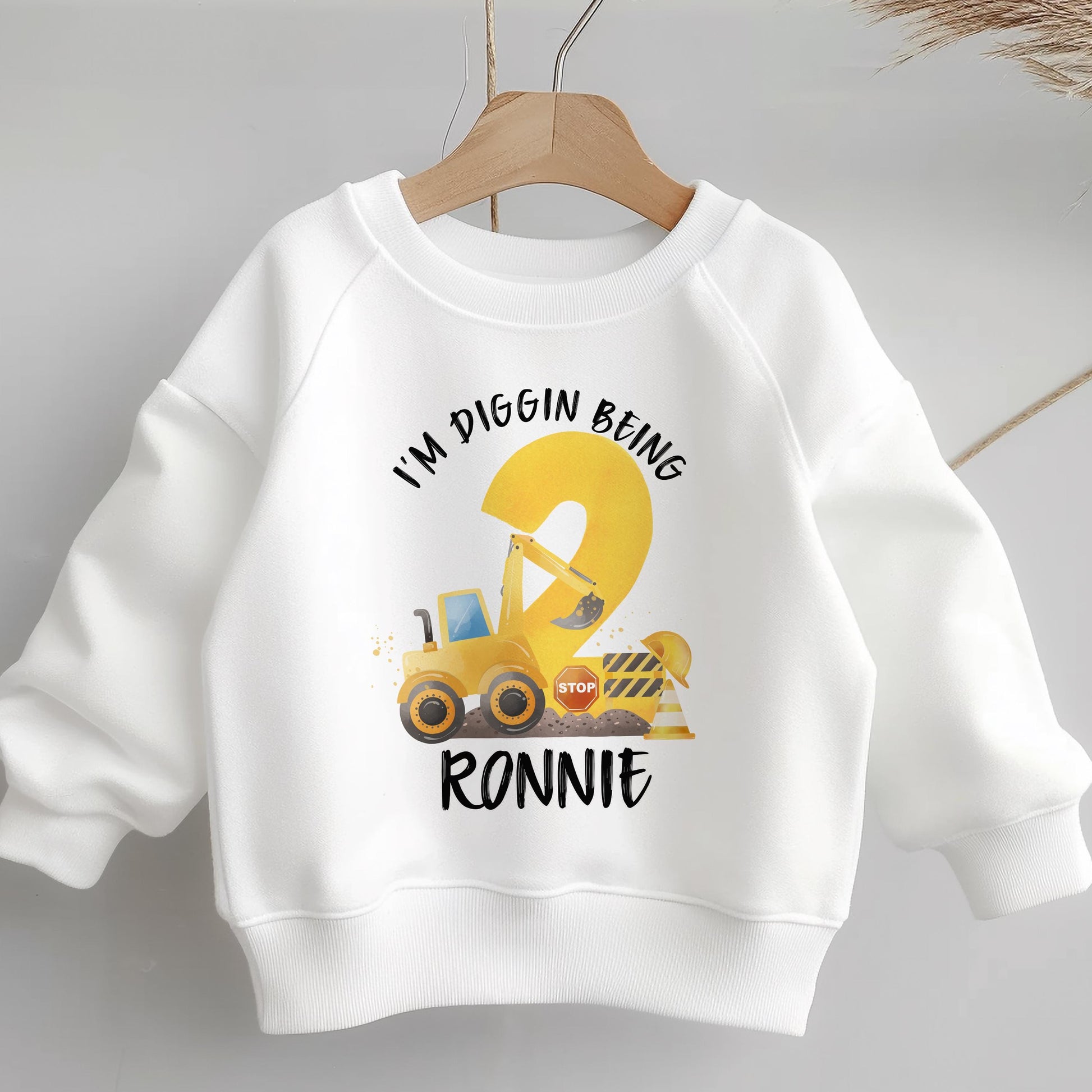 White children's sweatshirt on a hanger printed with a yellow digger construction birthday design reading I'M DIGGIN BEING