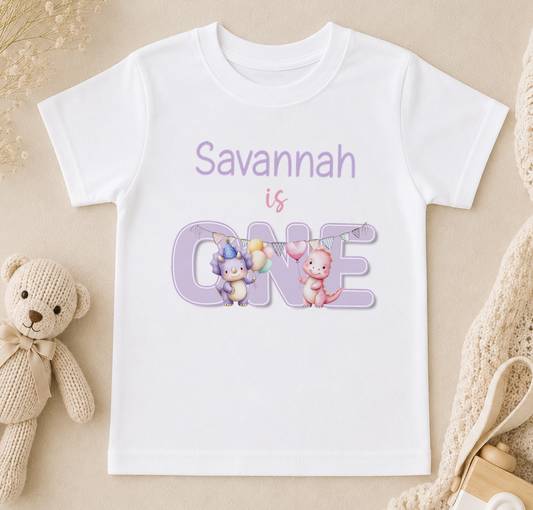 A white t-shirt with a graphic design featuring two cartoon animals, one pink and one blue, along with the text "Savannah is ONE" in purple.