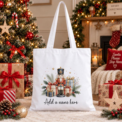 White cotton tote bag printed with a watercolour nutcracker toy soldier among wrapped presents and pine, above a name in script.