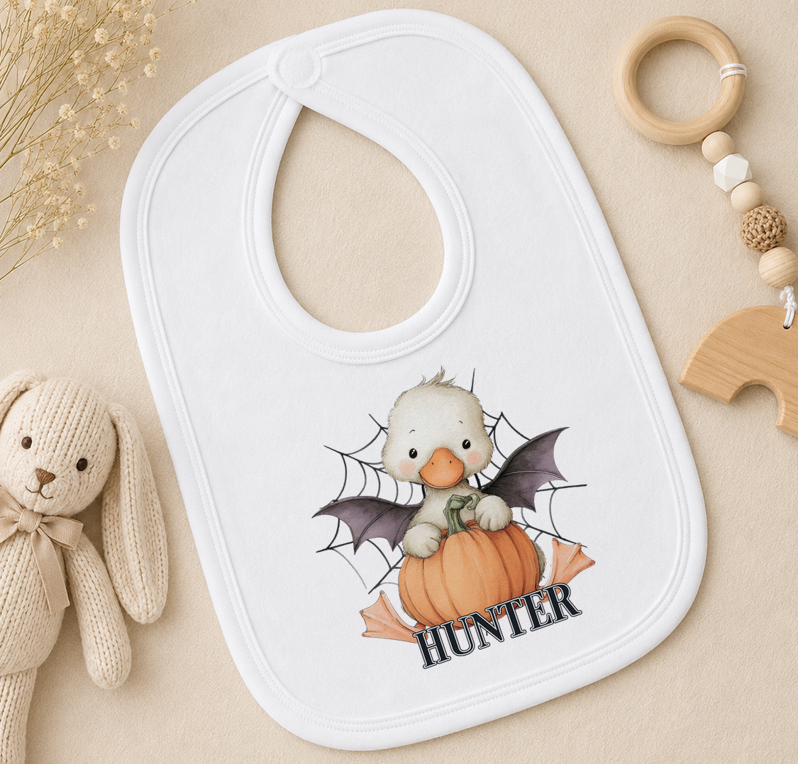A white bib with a cartoon image of a duck holding a pumpkin and the word "Hunter" printed on it, along with a wooden toy and a beaded ringer.