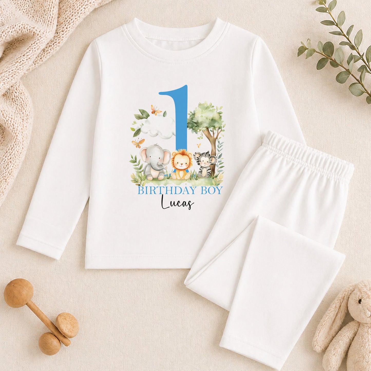 A white long-sleeved shirt with a blue number 1 and a graphic of animals, along with a pair of white pants, are displayed on a beige surface. There are also some toys and greenery in the background.