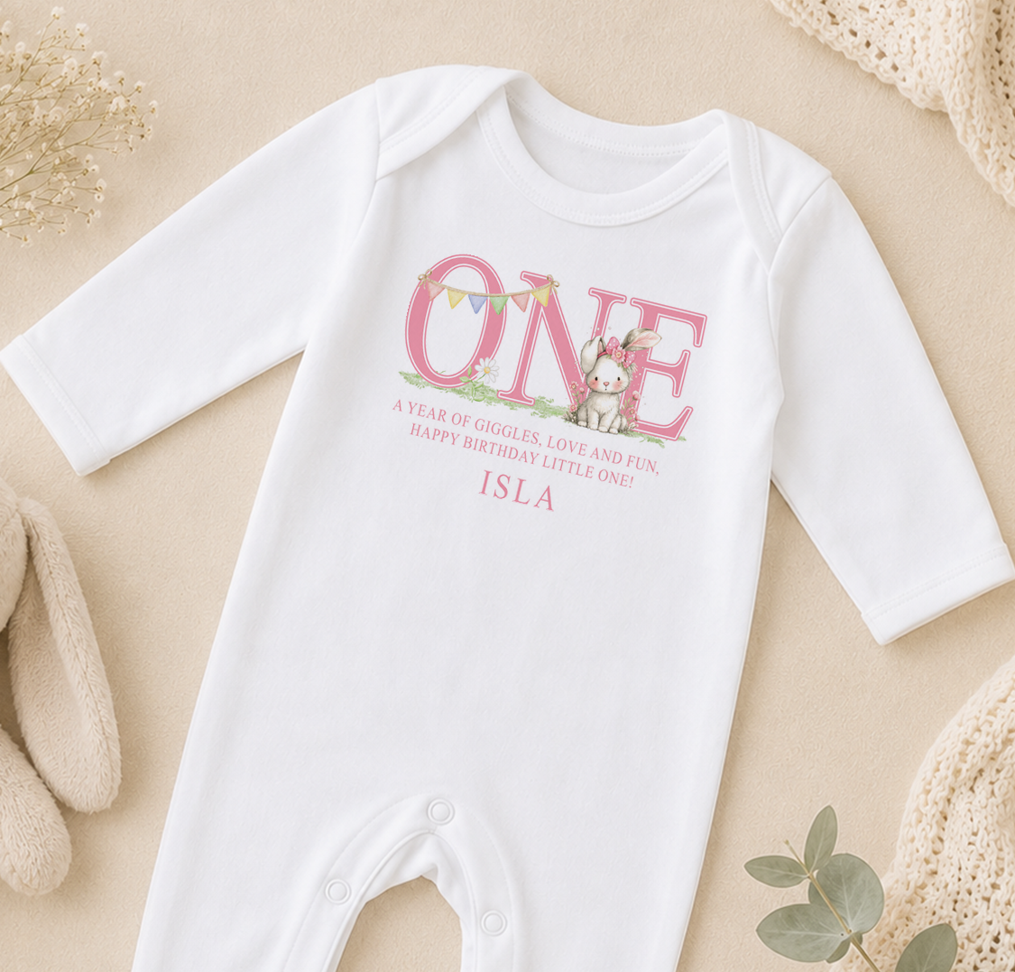 A white onesie with a pink bunny and the text "ONE" printed on it, along with a birthday message.