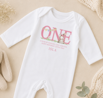 A white onesie with a pink bunny and the text "ONE" printed on it, along with a birthday message.