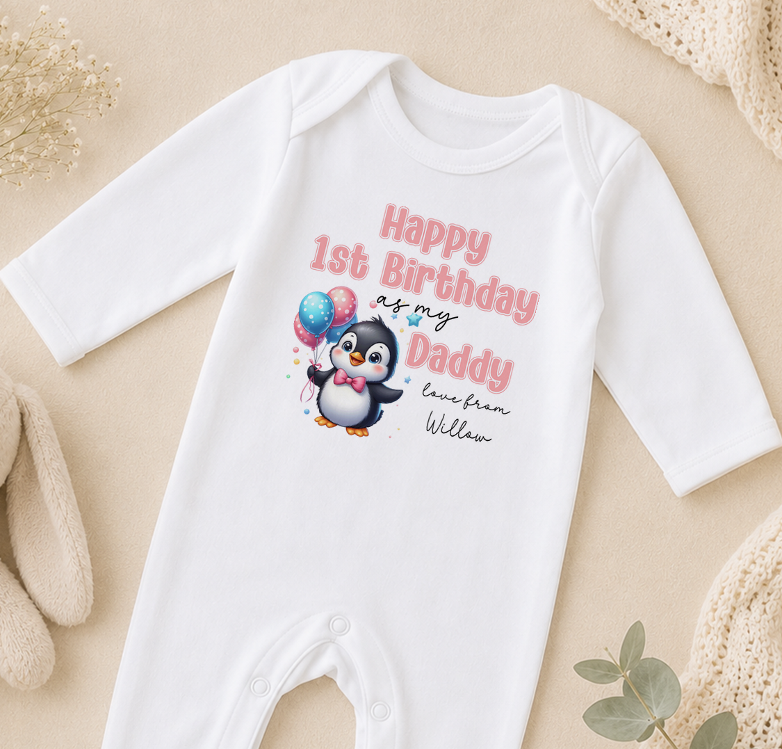 A white onesie with a penguin graphic and text that reads "Happy 1st Birthday as my Daddy love from William" is displayed on a beige surface.