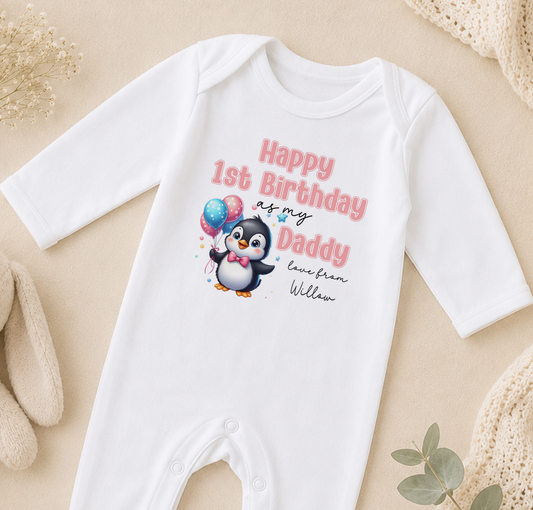 A white onesie with a penguin graphic and text that reads "Happy 1st Birthday as my Daddy love from William" is displayed on a beige surface.
