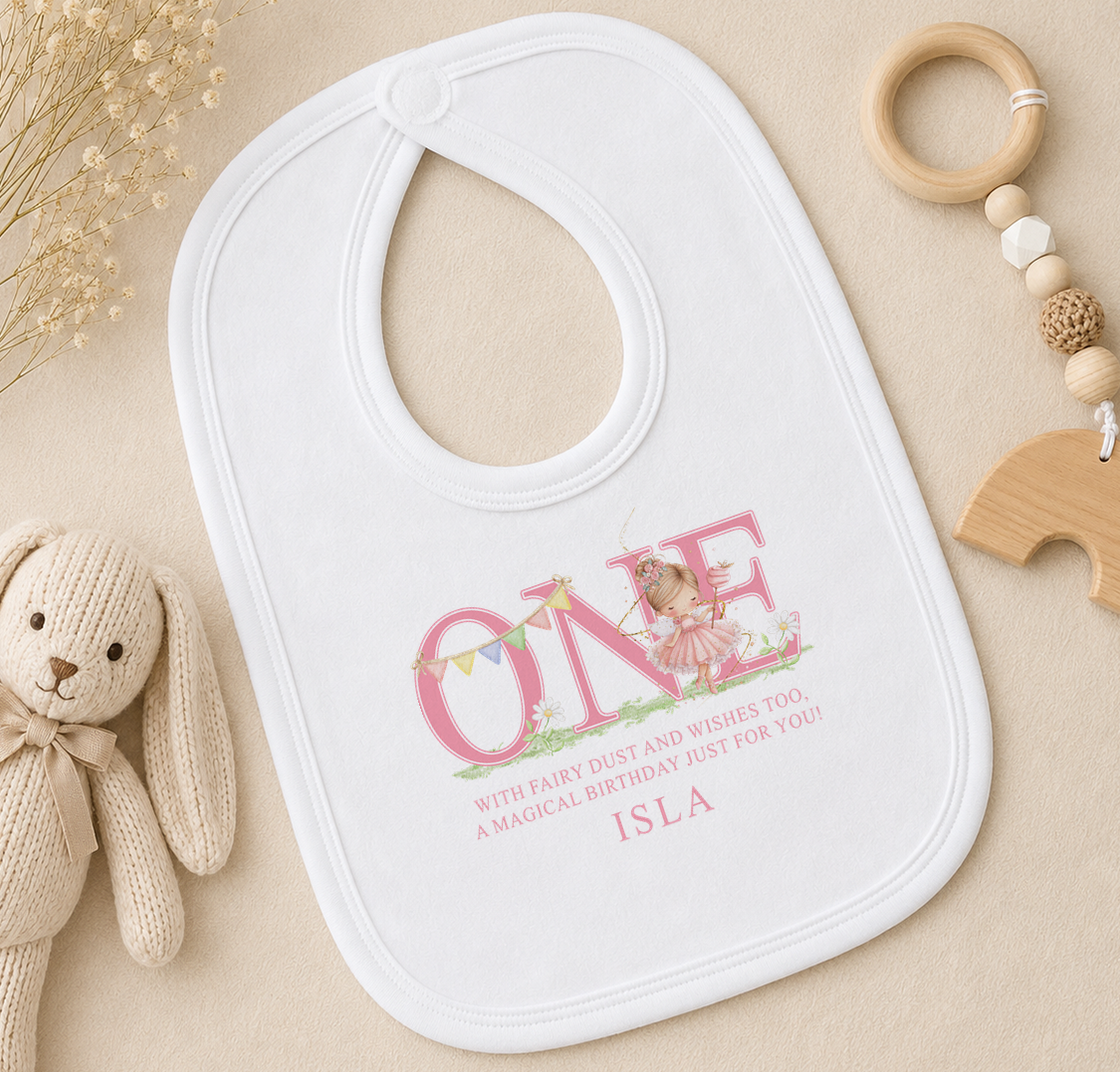 A white bib with a personalized message, a wooden toy, and some dried flowers on a beige surface.