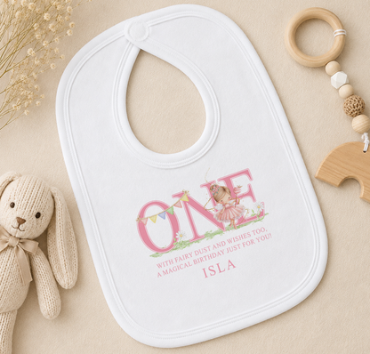 A white bib with a personalized message, a wooden toy, and some dried flowers on a beige surface.