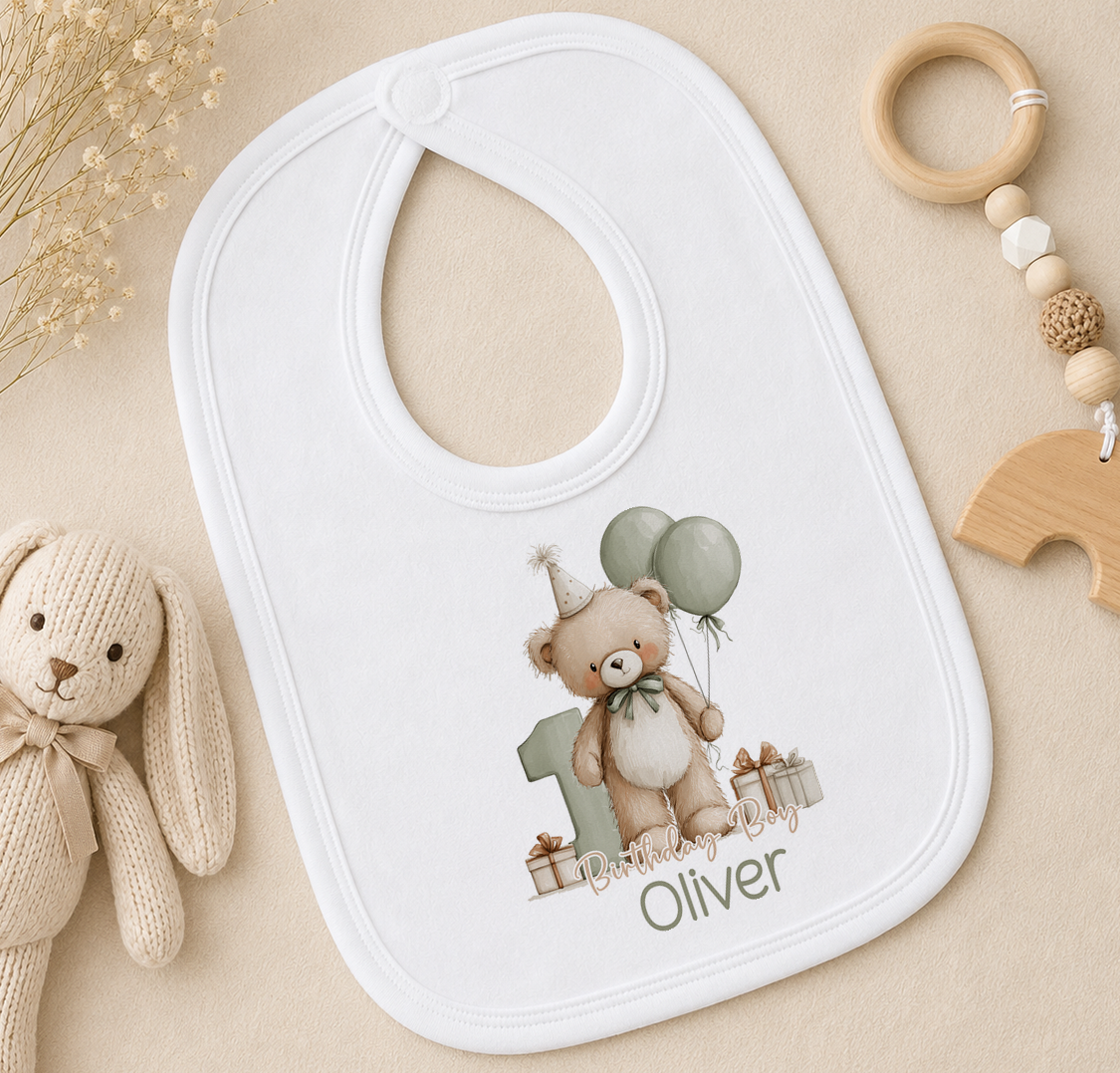A white bib with a teddy bear and balloons printed on it, along with a wooden toy and a wooden bead ring, all placed on a beige surface.