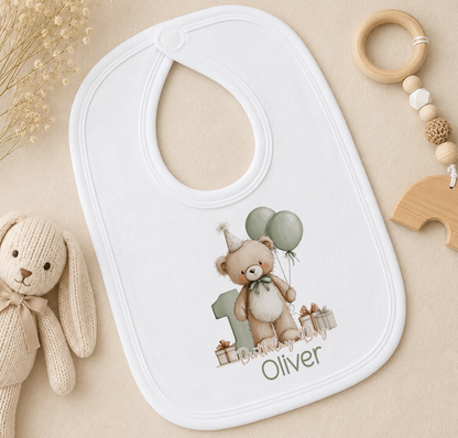 A white bib with a teddy bear and balloons printed on it, along with a wooden toy and a wooden bead ring, all placed on a beige surface.
