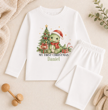 A white long-sleeved shirt with a Christmas-themed graphic of a green dinosaur wearing a Santa hat and scarf, along with the text "My first Christmas Daniel". The shirt is placed on a beige background, and there are some Christmas decorations and toys visible in the image.