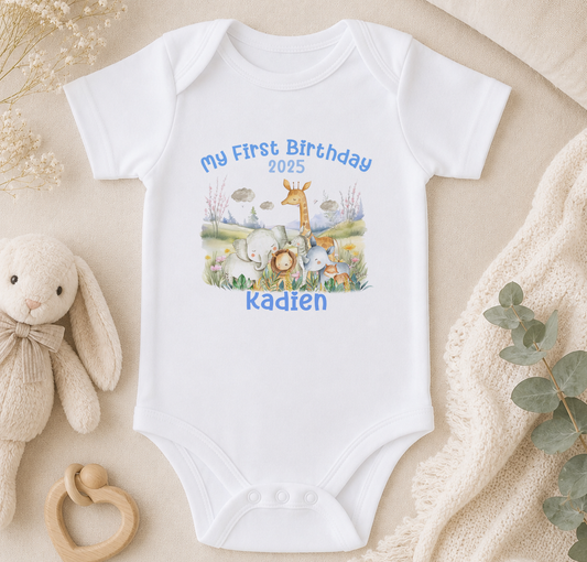 A white onesie with a printed image of animals and the text "My First Birthday 2020 Kadien" is displayed on a bed, accompanied by a stuffed animal and a beige blanket.
