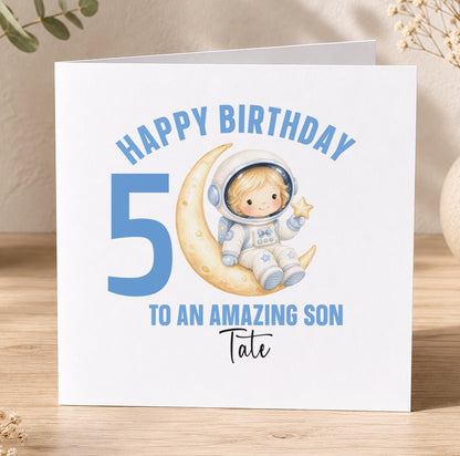 Personalised astronaut birthday card design showing a cartoon spaceman floating on a crescent moon among stars, with space for a child's personalised name and age.