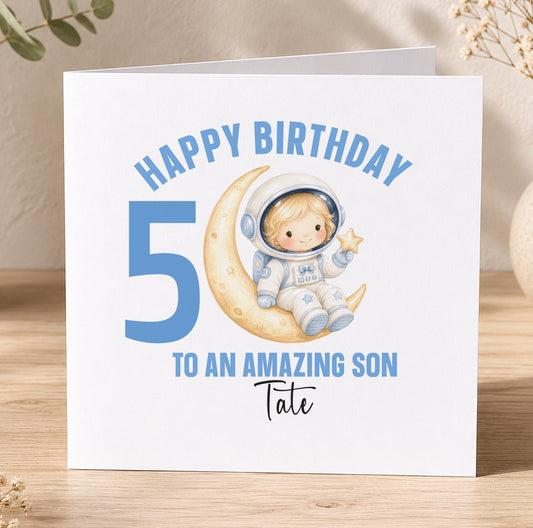 Personalised astronaut birthday card design showing a cartoon spaceman floating on a crescent moon among stars, with space for a child's personalised name and age.