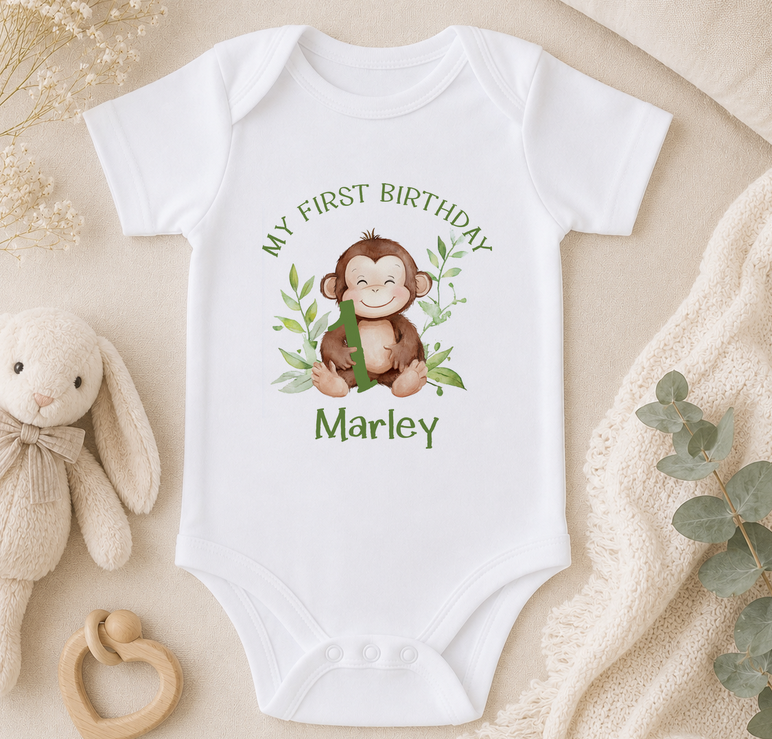 A white onesie with a monkey graphic and the text "My first birthday Marley" printed on it, along with a stuffed animal and some greenery.