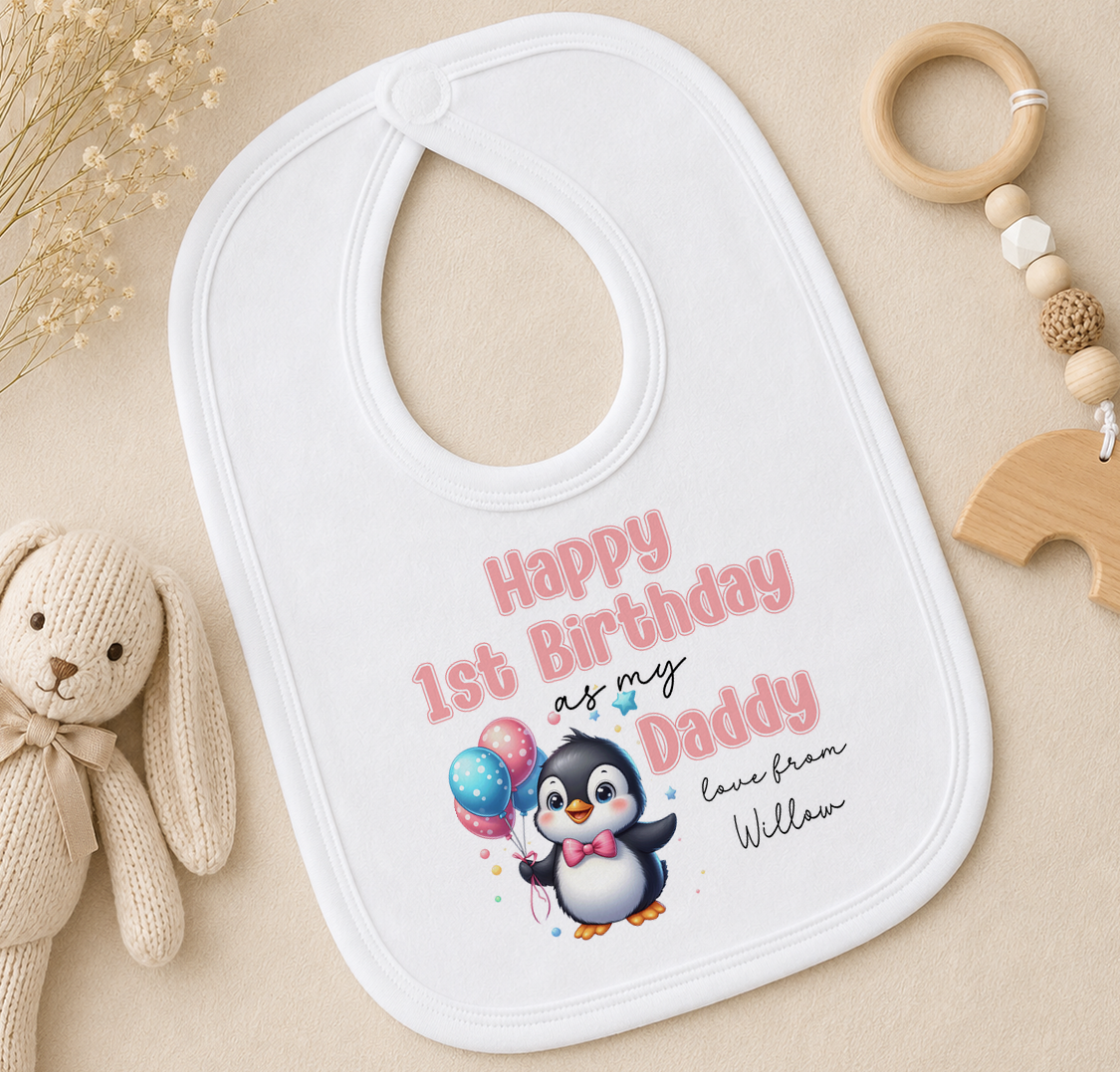 A white baby bib with a penguin graphic and text, along with a wooden toy and a beaded ring, arranged on a beige surface.