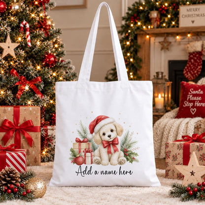 White cotton tote bag printed with a cream puppy in a red Santa hat sitting beside wrapped gifts, baubles and holly, above a name in script.