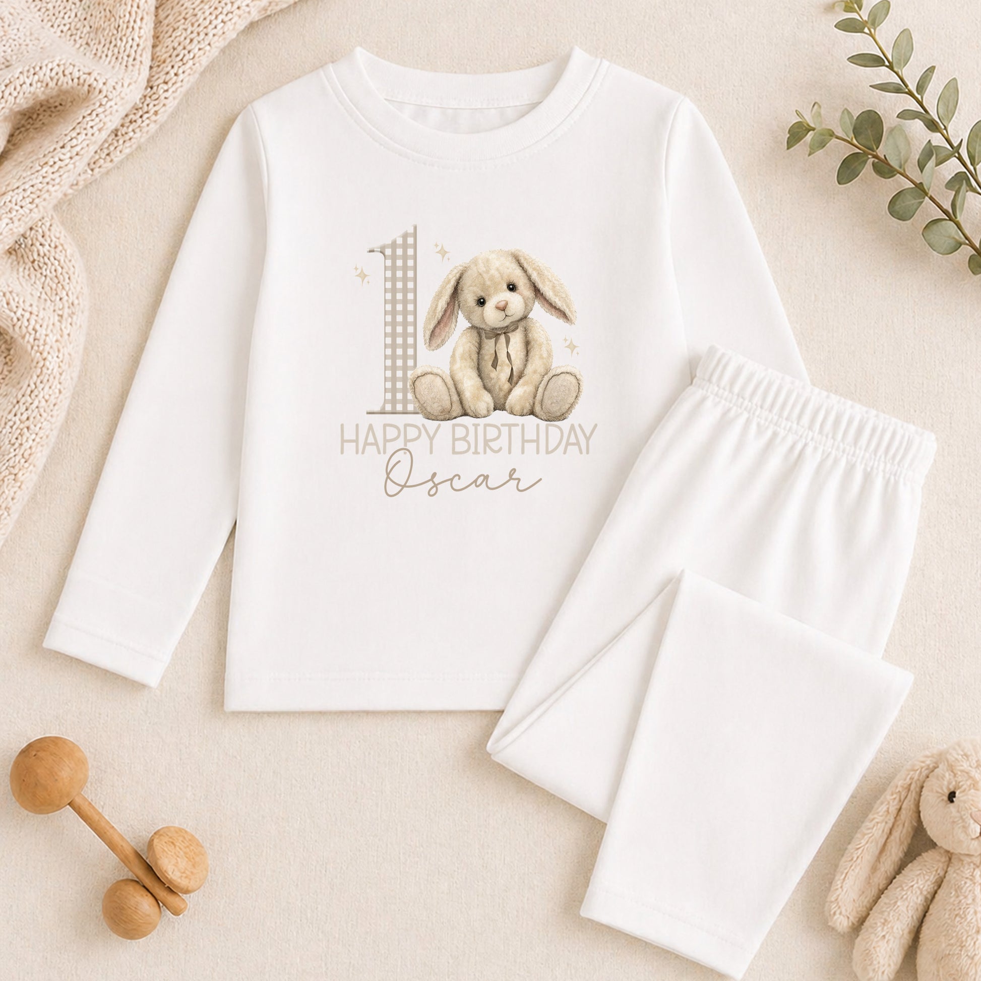 A white onesie with a bunny graphic and the text "Happy Birthday Oscar" printed on it, along with a pair of white pajama pants and a stuffed animal toy.