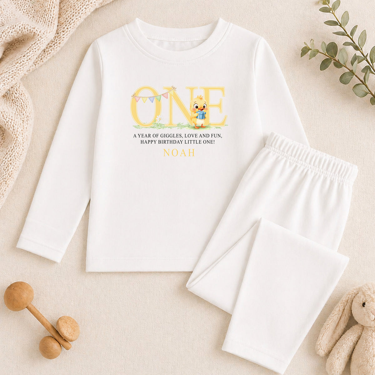 A white long-sleeved shirt with a personalized message and a small cartoon character printed on it, along with a pair of white pants and some toys.