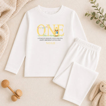 A white long-sleeved shirt with a personalized message and a small cartoon character printed on it, along with a pair of white pants and some toys.