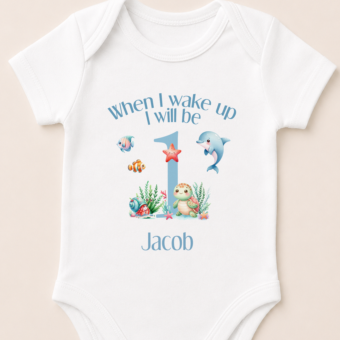A white onesie with a personalized design for a baby's first birthday. The design includes a blue number 1, a dolphin, a starfish, and other marine life. The text above the design says "When I wake up I will be Jacob".
