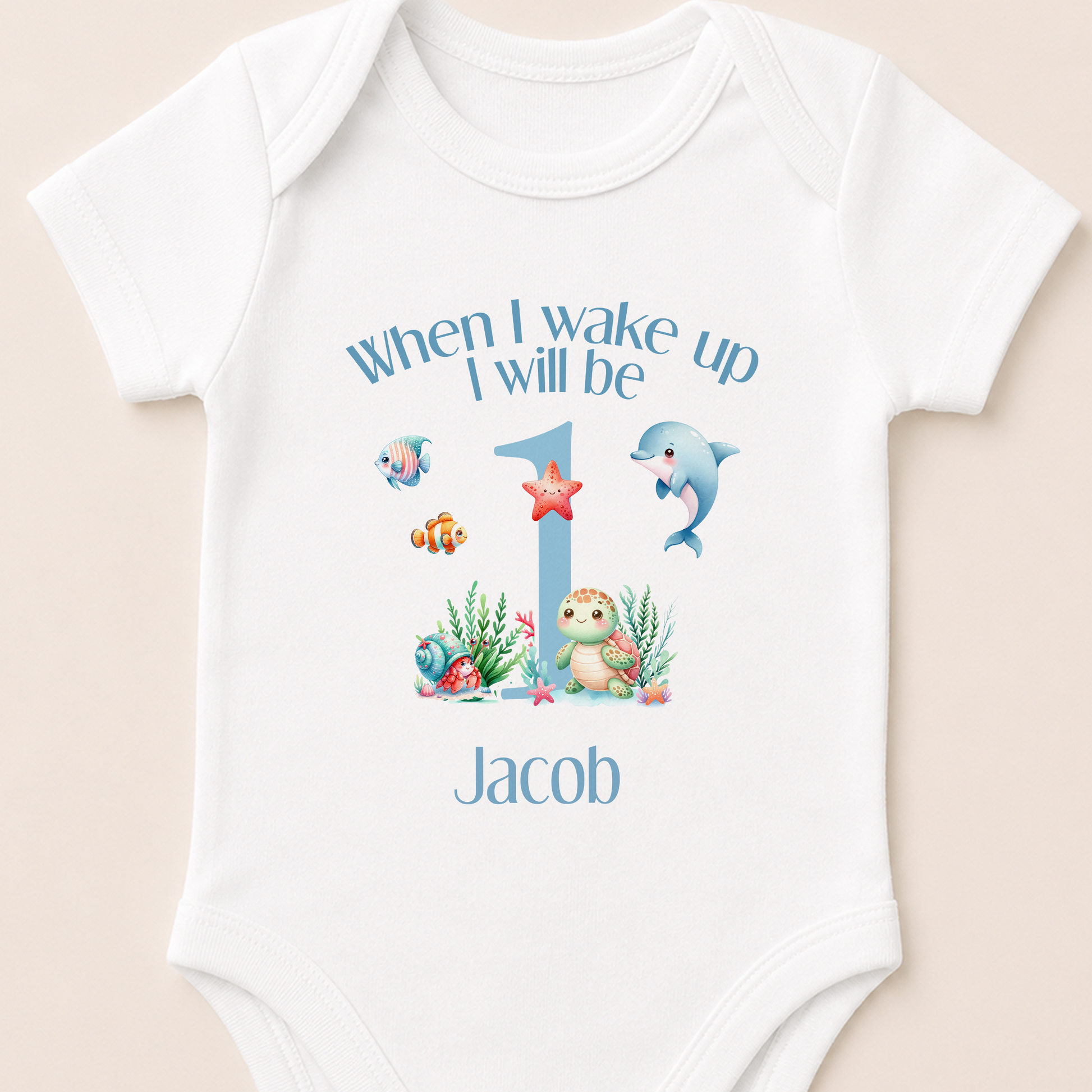 A white onesie with a personalized design for a baby's first birthday. The design includes a blue number 1, a dolphin, a starfish, and other marine life. The text above the design says "When I wake up I will be Jacob".