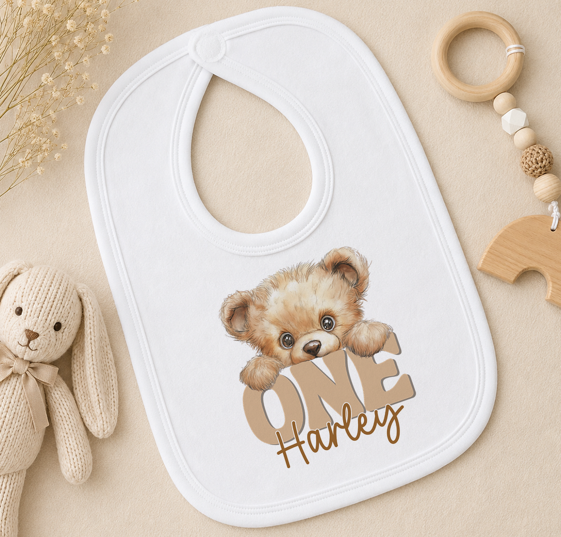 A white bib with a teddy bear design and the word "Harley" printed on it, along with a wooden toy and a wooden bead ring.