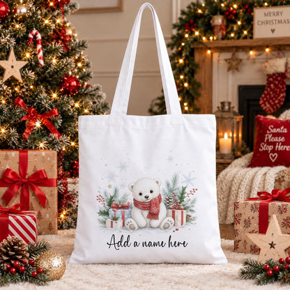 White cotton tote bag printed with a polar bear cub in a red scarf beside wrapped gifts, pine and snowflakes, above a name in script.