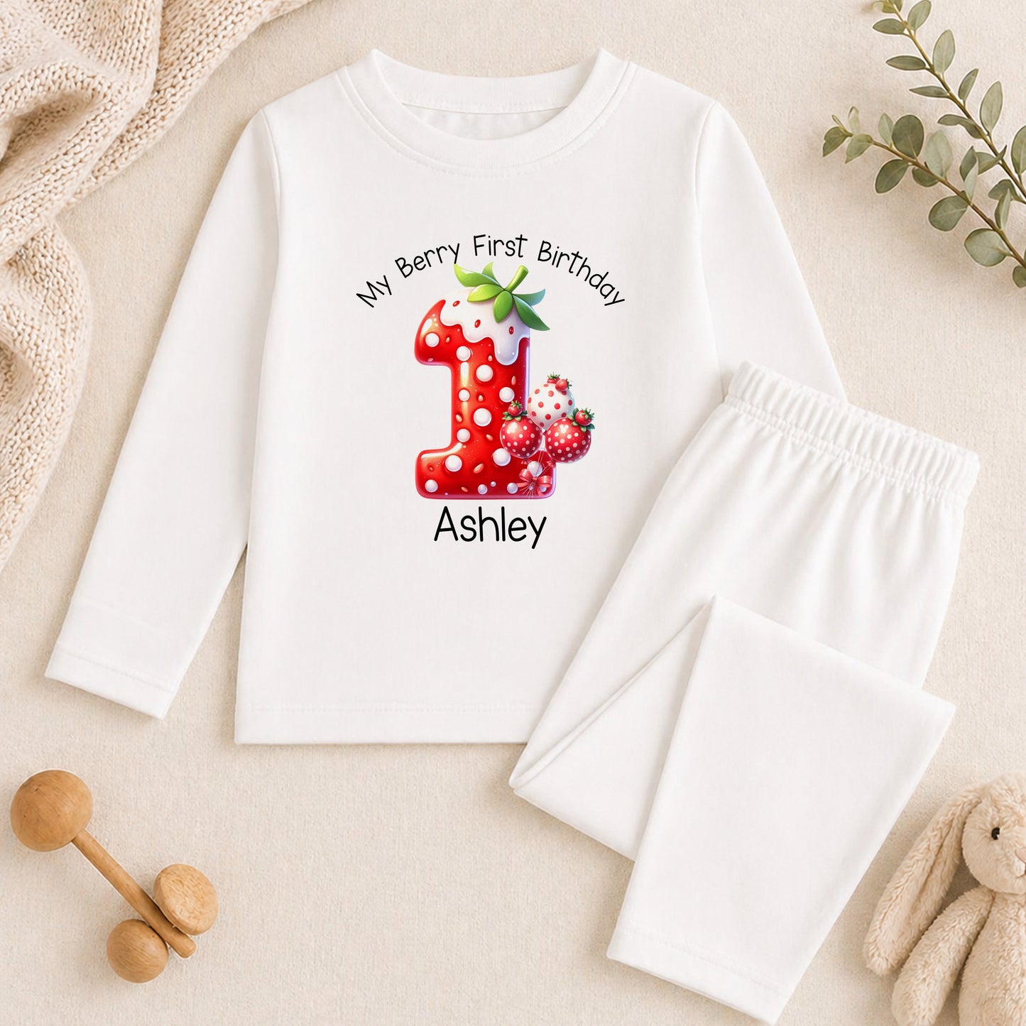 A white long-sleeved shirt with a personalized number one and strawberry design, along with a pair of white pants, a stuffed animal, and some green leaves.