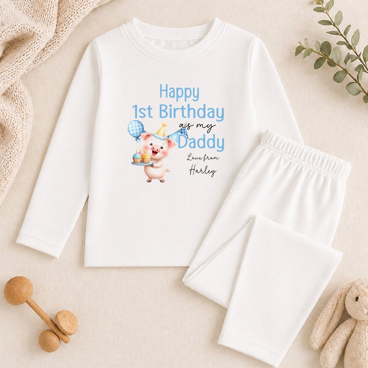 A white long-sleeved shirt with a cartoon pig and text that says "Happy 1st Birthday is my Daddy Love from Harley" is displayed on a beige surface, along with a pair of white pants and a stuffed animal.