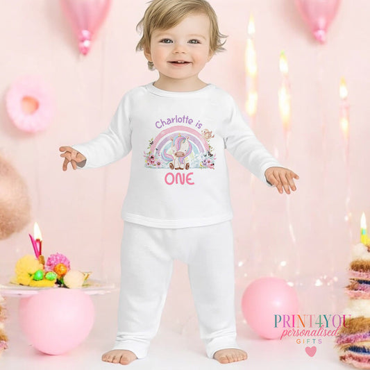 Personalised unicorn and rainbow first birthday pyjamas with the baby's name and 'I am ONE' printed on soft cotton, 6-12 months size shown.