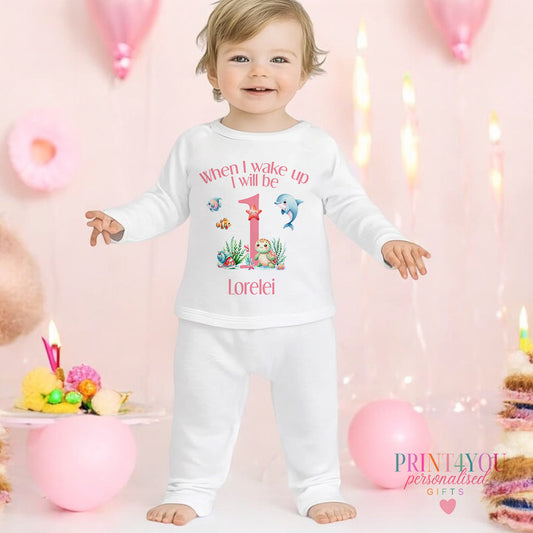 Personalised pink sea theme first birthday pyjamas with dolphins, starfish and the baby's name printed on soft cotton, 6-12 months size shown.