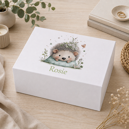 Personalised sage green baby hedgehog keepsake box with magnetic lid, showing a hedgehog wearing a floral crown resting on a green cushion with the name Rosie in script, on a wooden tabletop with eucalyptus and candles