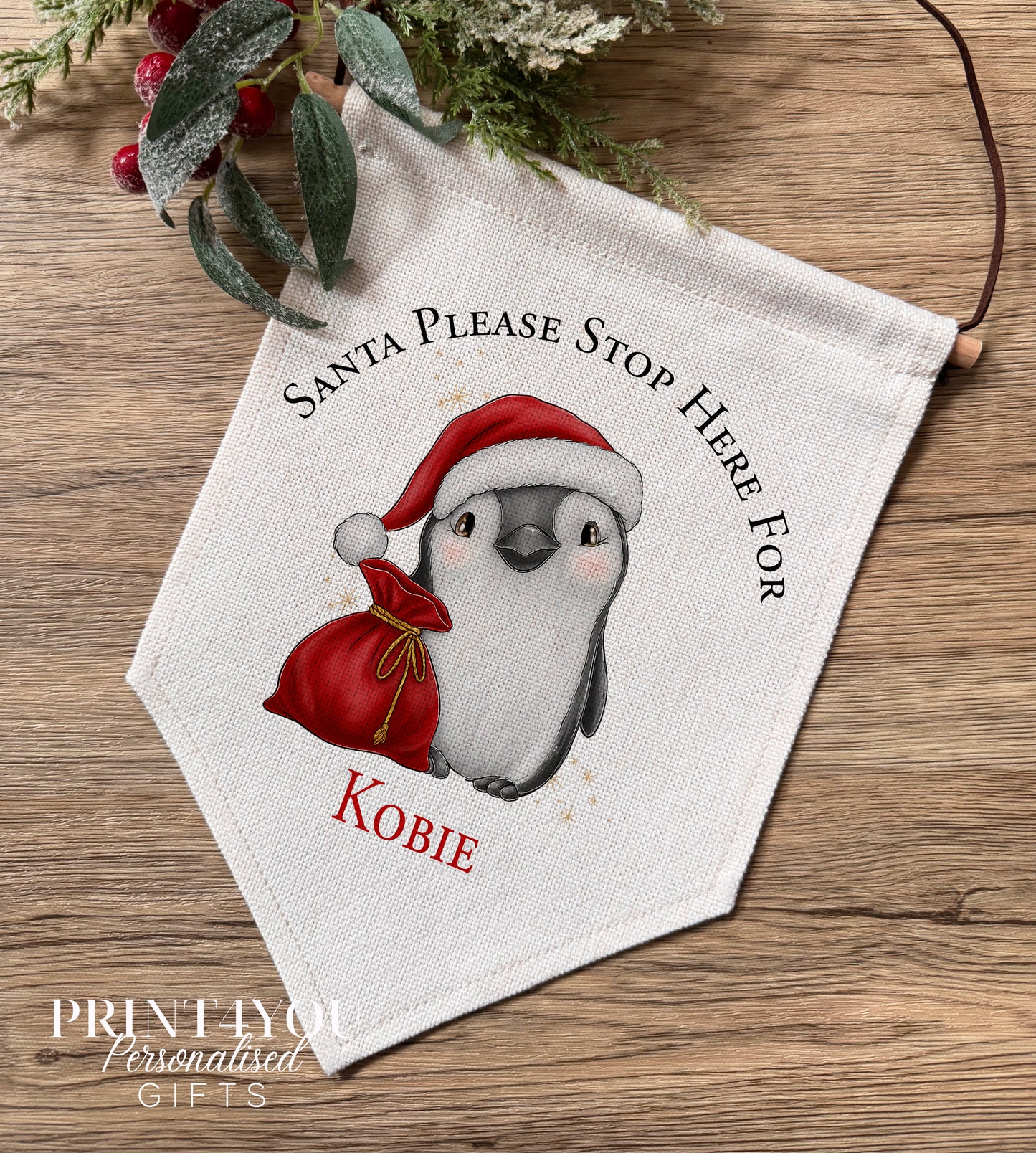 Personalised linen Christmas banner featuring a penguin wearing a Santa hat beside a Santa Stop Here message, personalised with the name Millie, hung on a wooden bar