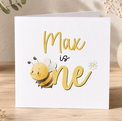 Personalised honey bee 1st birthday card with cartoon bee and floral design, custom name reads Is One